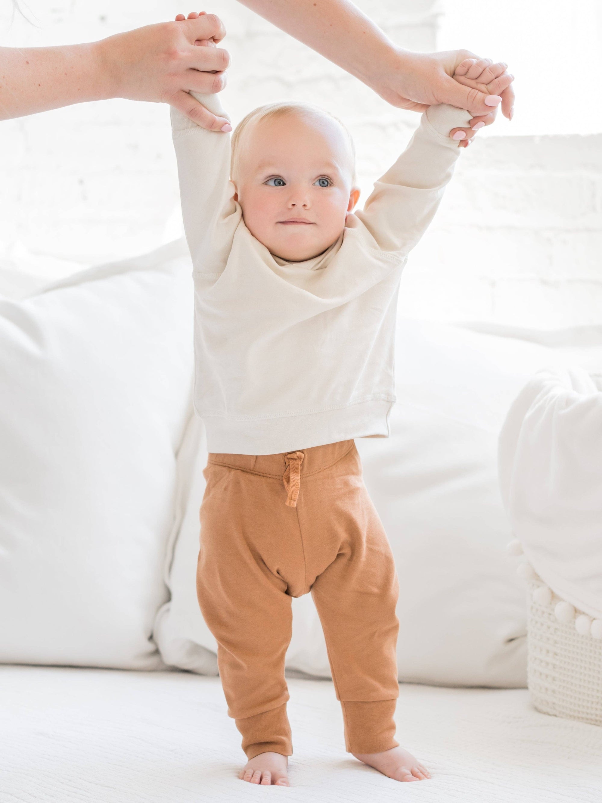 Baby standing with arms raised, wearing a white sweater and brown pants, on a white background.